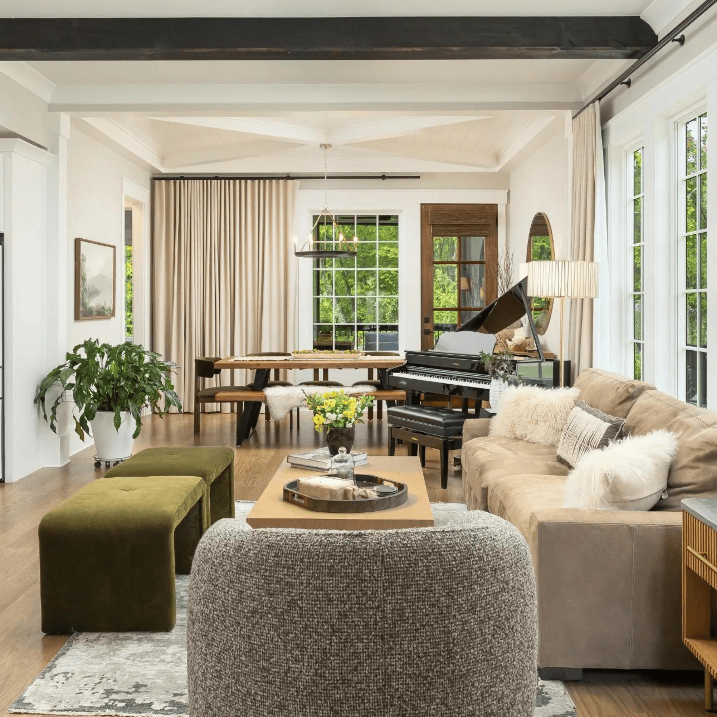 Spacious living room with a large window, plush seating, a piano, and wooden accents. The home boasts green ottomans, a plant, and a centerpiece on the coffee table. Natural light fills the room, highlighting the interior design that makes this Chattanooga residence truly inviting.