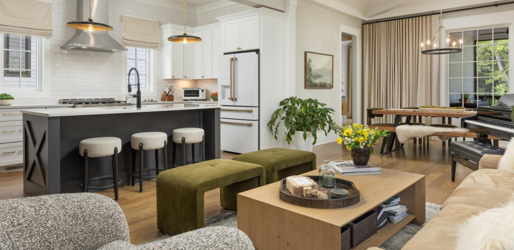 Open-concept living space featuring a kitchen with an island, bar stools, and stainless steel appliances, adjacent to a living room with a coffee table, green benches, and a dining area with modern lighting.