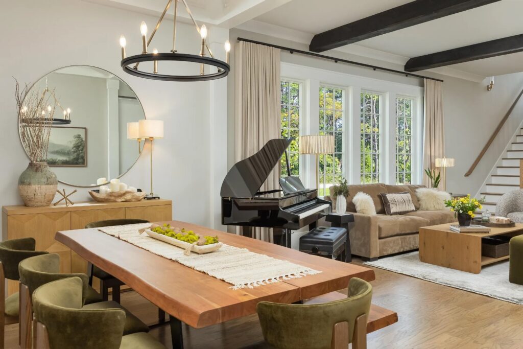 A modern living and dining room features a wooden table with green chairs, a beige sofa, a grand piano, large windows, and neutral decor with black ceiling beams.