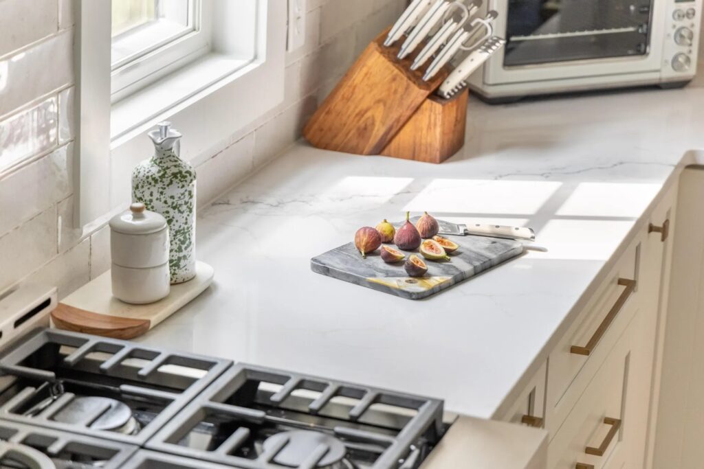 A marble cutting board with sliced figs and a knife sits on a white kitchen countertop near a stove, window, and knife block.
