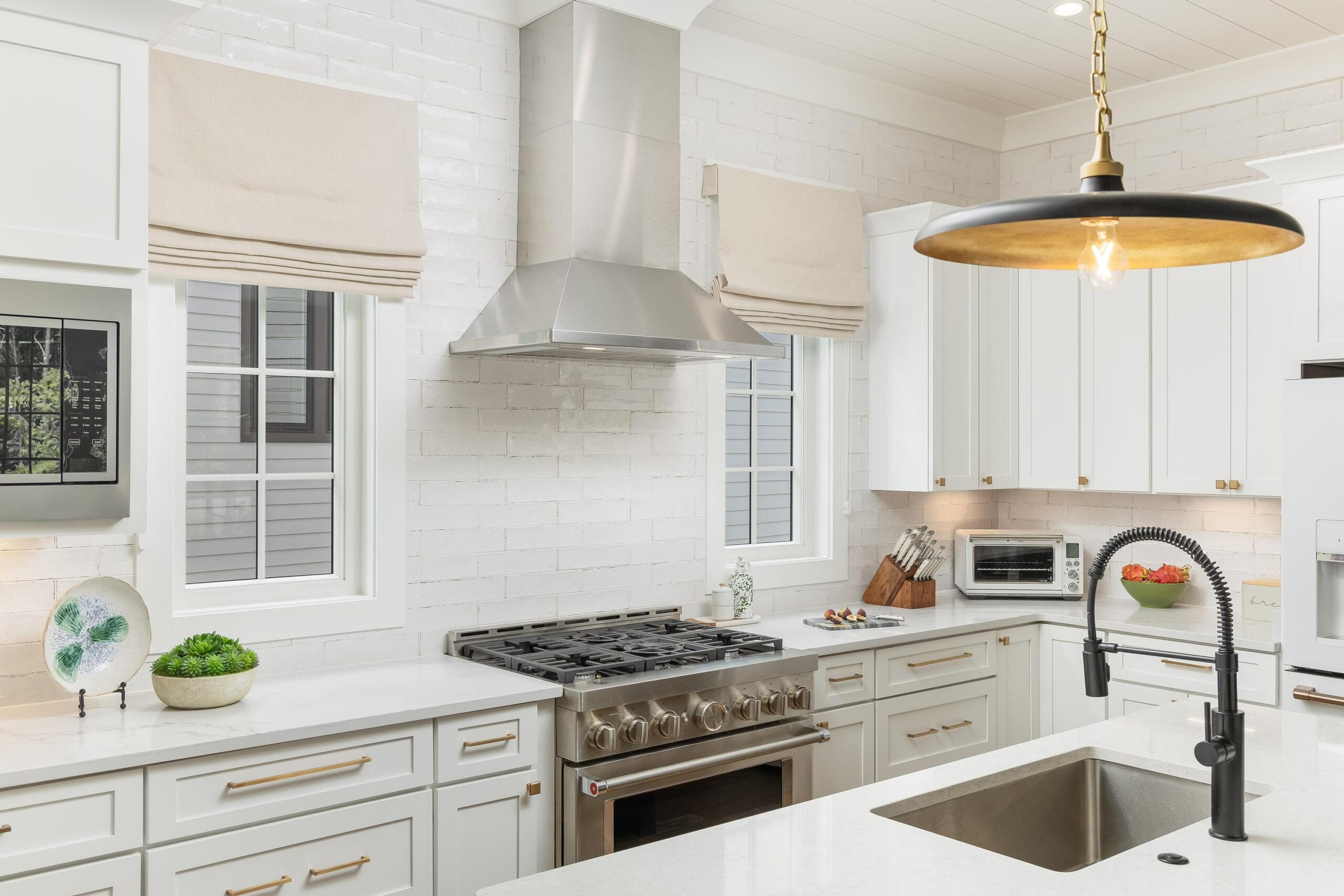 Modern, full-service kitchen featuring white cabinets, stainless steel stove and range hood, farmhouse sink, pendant light, and two windows with beige Roman shades.
