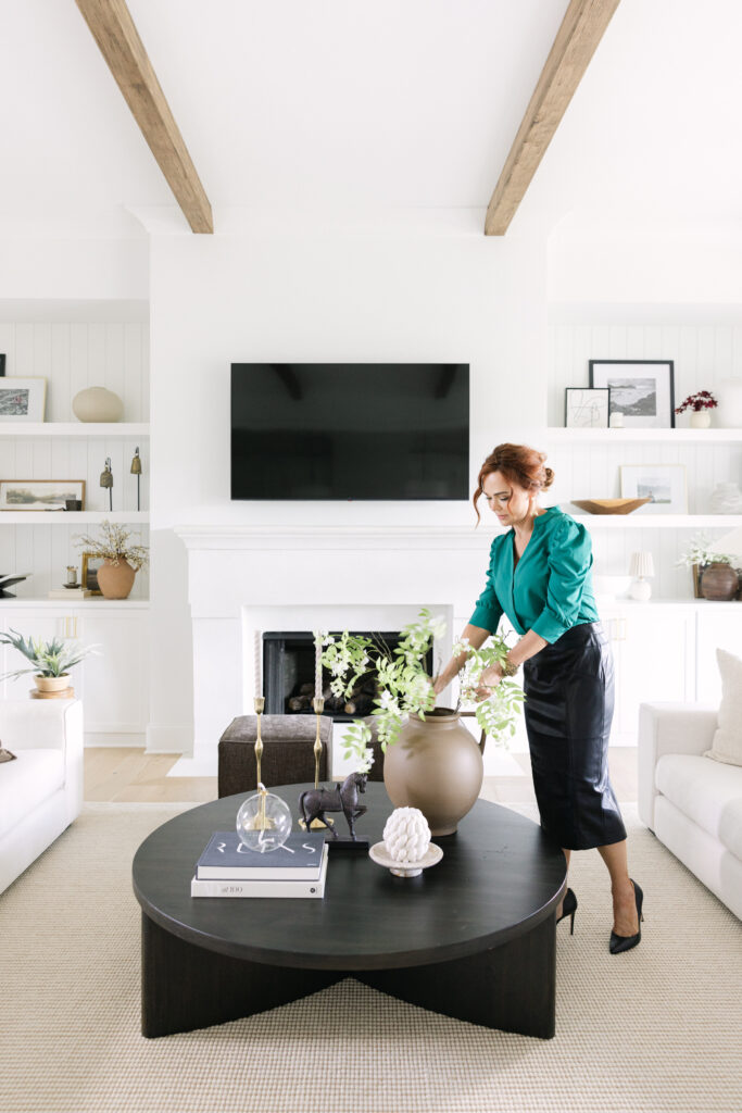A woman in a green top arranges branches in a vase on a black coffee table, adding designer touches to a bright, modern living room with a fireplace and built-in shelves.