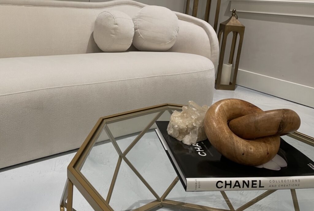 A designer geometric glass coffee table with a Chanel book, crystal, and wooden sculpture sits in front of a cream-colored sofa with round cushions.