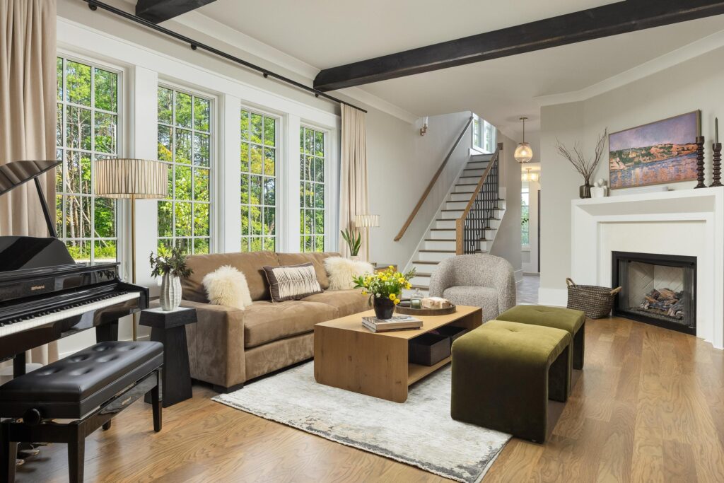 A modern living room perfect for hosting, featuring large windows, a brown sofa, two ottomans, a piano, a wooden coffee table, a fireplace, and a staircase in the background.