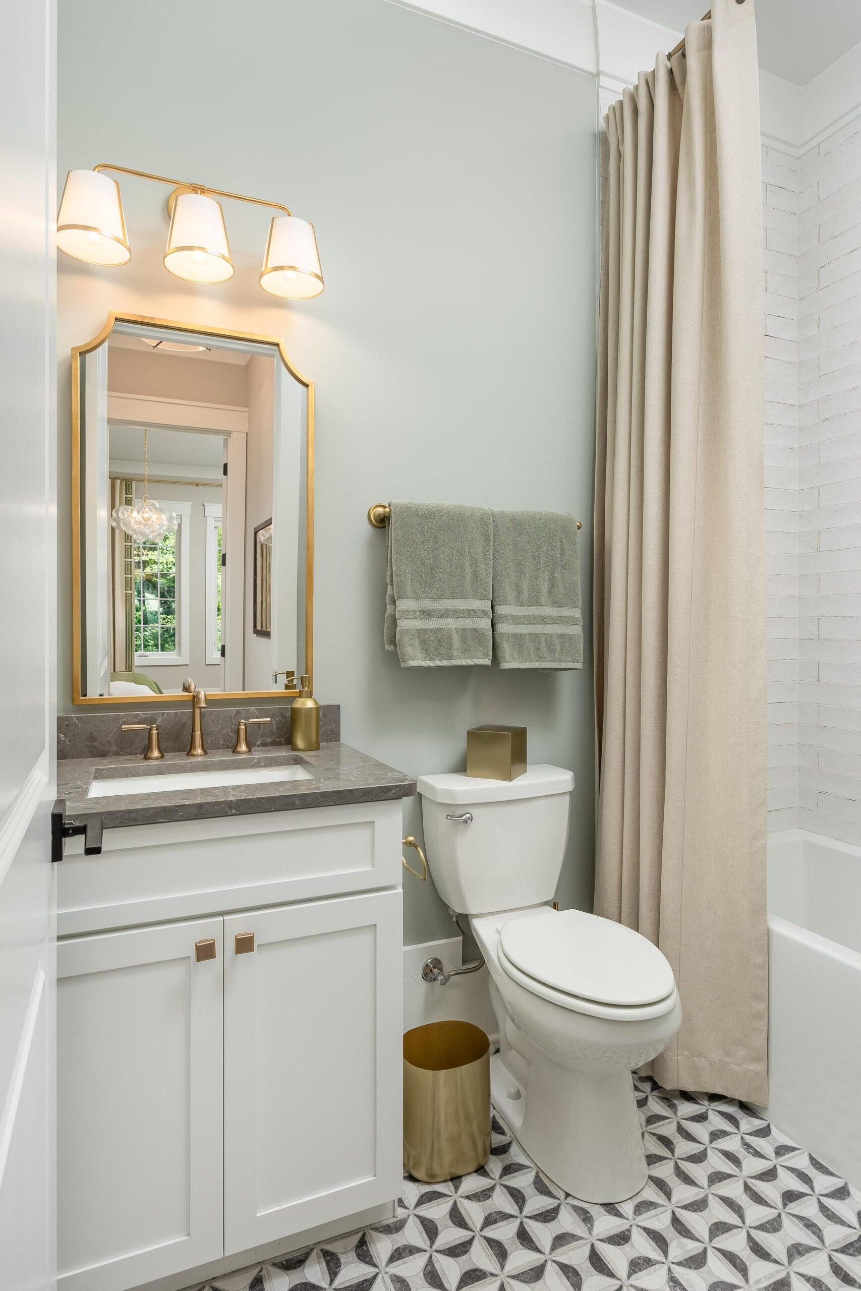 Modern bathroom with a white vanity, gold fixtures, mirror, towel rack with two green towels, toilet, gold trash can, patterned tile floor, and shower with beige curtain and white tile.