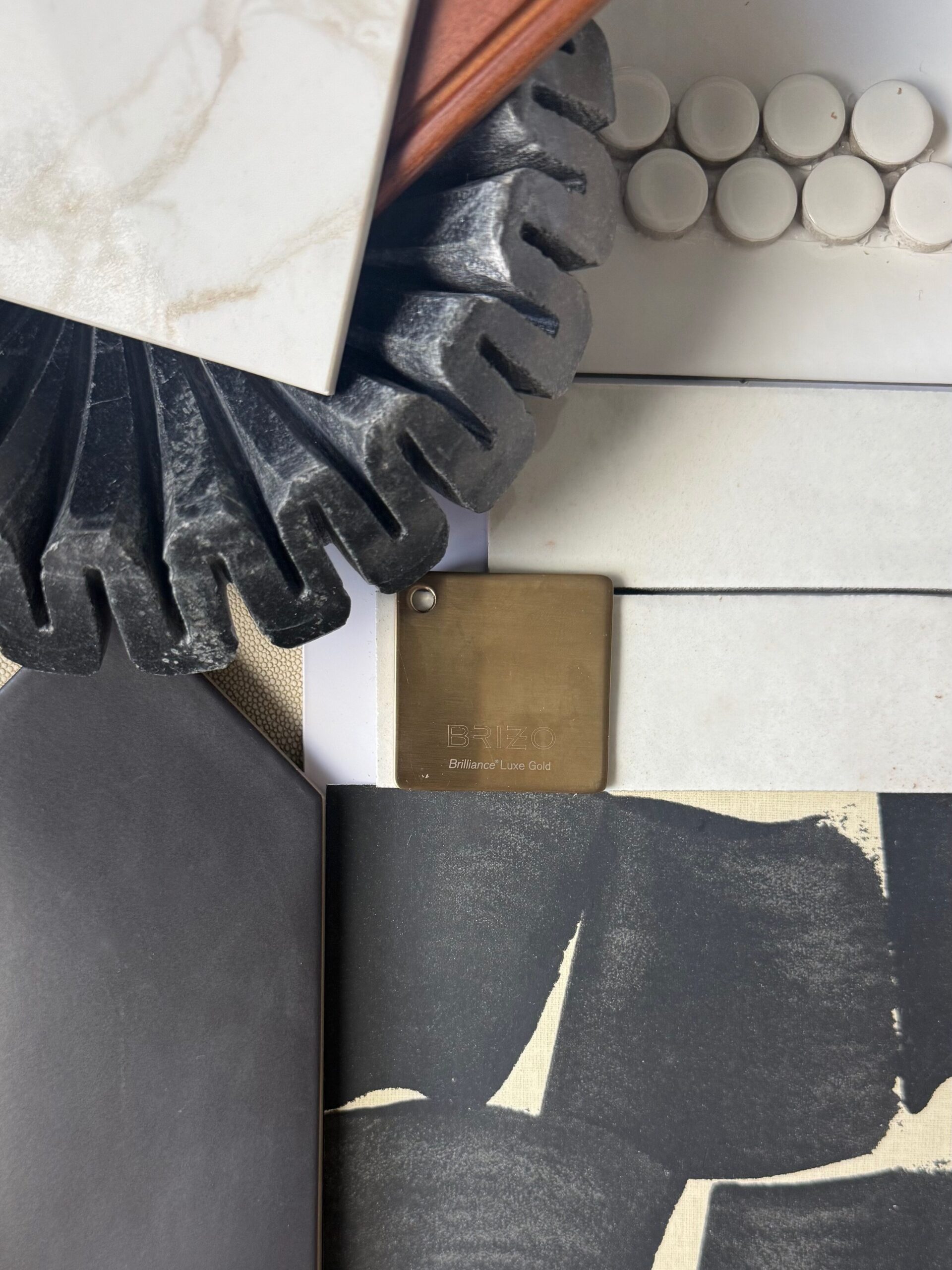 A flat lay of various interior design materials, including tile samples, a black textured object, white round tiles, a large black tile, a gold metal sample, and a black patterned fabric.