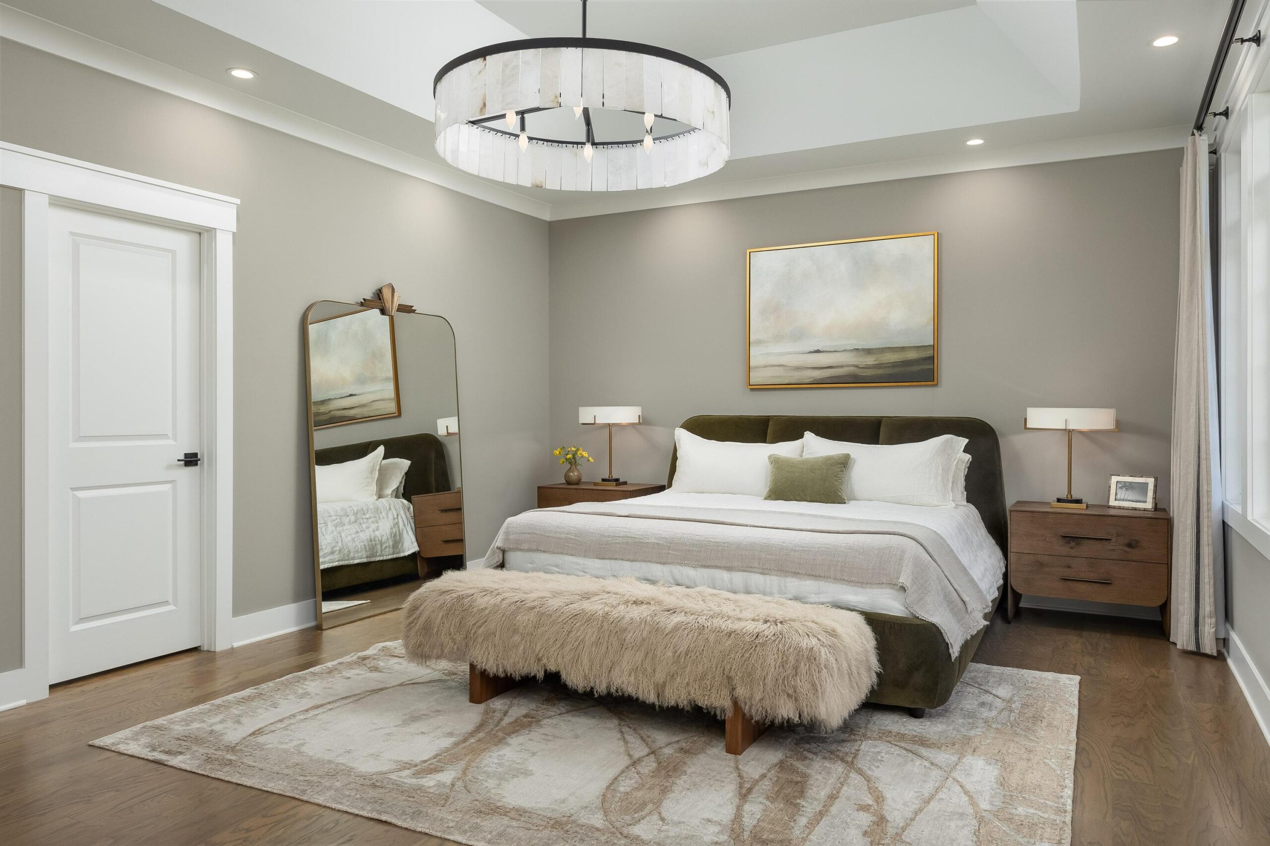 Modern bedroom with a large bed, wooden nightstands, a fuzzy bench, tile flooring, a floor mirror, a landscape painting above the bed, and a round ceiling light. Neutral colors and minimal decor.
