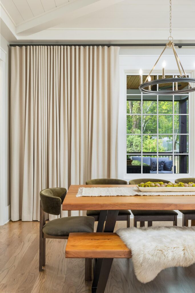 Modern dining room with a wooden table, green velvet chairs, a bench with a sheepskin throw, beige curtains, and large windows showing greenery outside. Sleek tile flooring adds a contemporary touch to the space.
