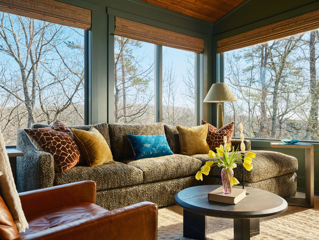 A cozy living room with a textured sofa, assorted pillows, and a round coffee table with a vase of yellow tulips creates the perfect space to showcase your interior design portfolio by large windows overlooking trees.