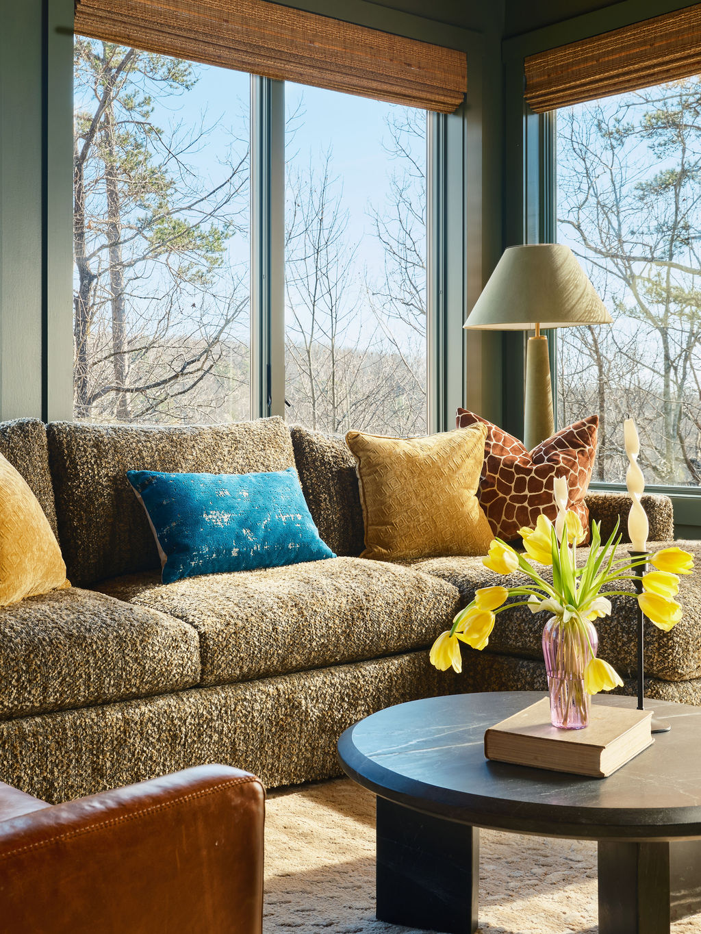 Sunlit living room with a textured sofa, colorful throw pillows, and a round black coffee table featuring yellow flowers in a vase—perfect for adding style to your portfolio. Large windows offer inviting views of trees outside.