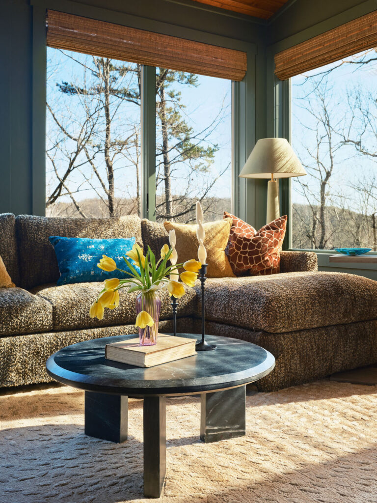 A sunlit living room with large windows, a patterned sofa with colorful pillows, and a round black coffee table holding a vase of yellow tulips, books, and two candlesticks—perfect for any interior design portfolio.