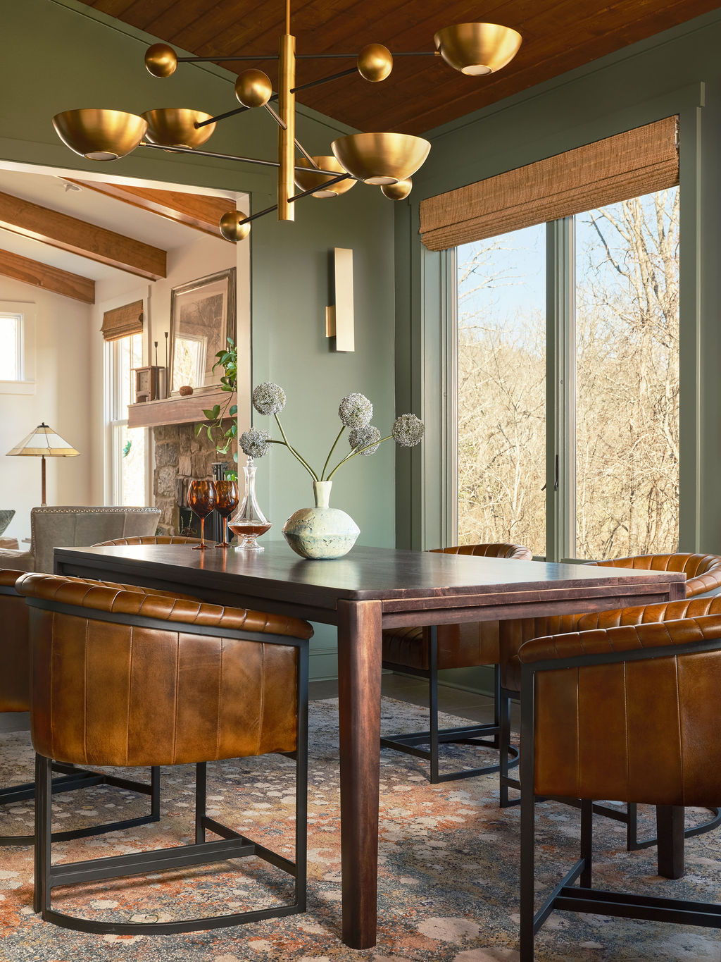 A modern dining room perfect for a design portfolio, featuring a dark wood table, four brown leather chairs, a gold chandelier, and large windows with an outdoor view. A ceramic vase with flowers sits on the table.