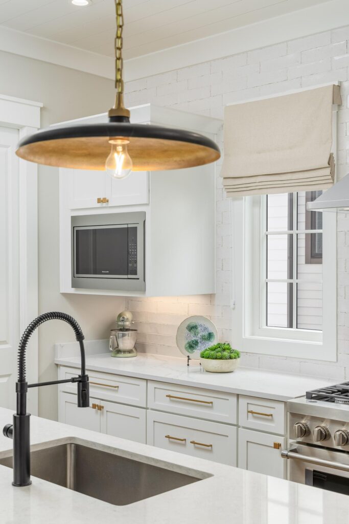 Modern white kitchen with brass hardware, a black and gold pendant light, a farmhouse sink, and a window with a beige Roman shade create a timeless kitchen ambiance.