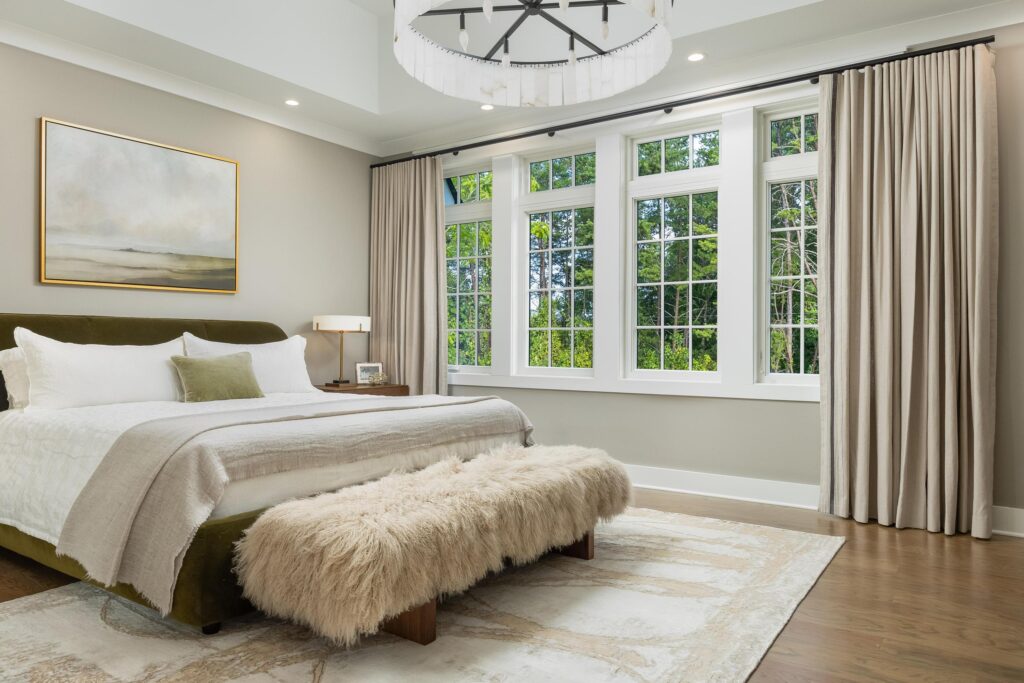 A modern bedroom with a large bed, a fuzzy bench, a landscape painting, beige window treatments, and tall windows overlooking greenery.