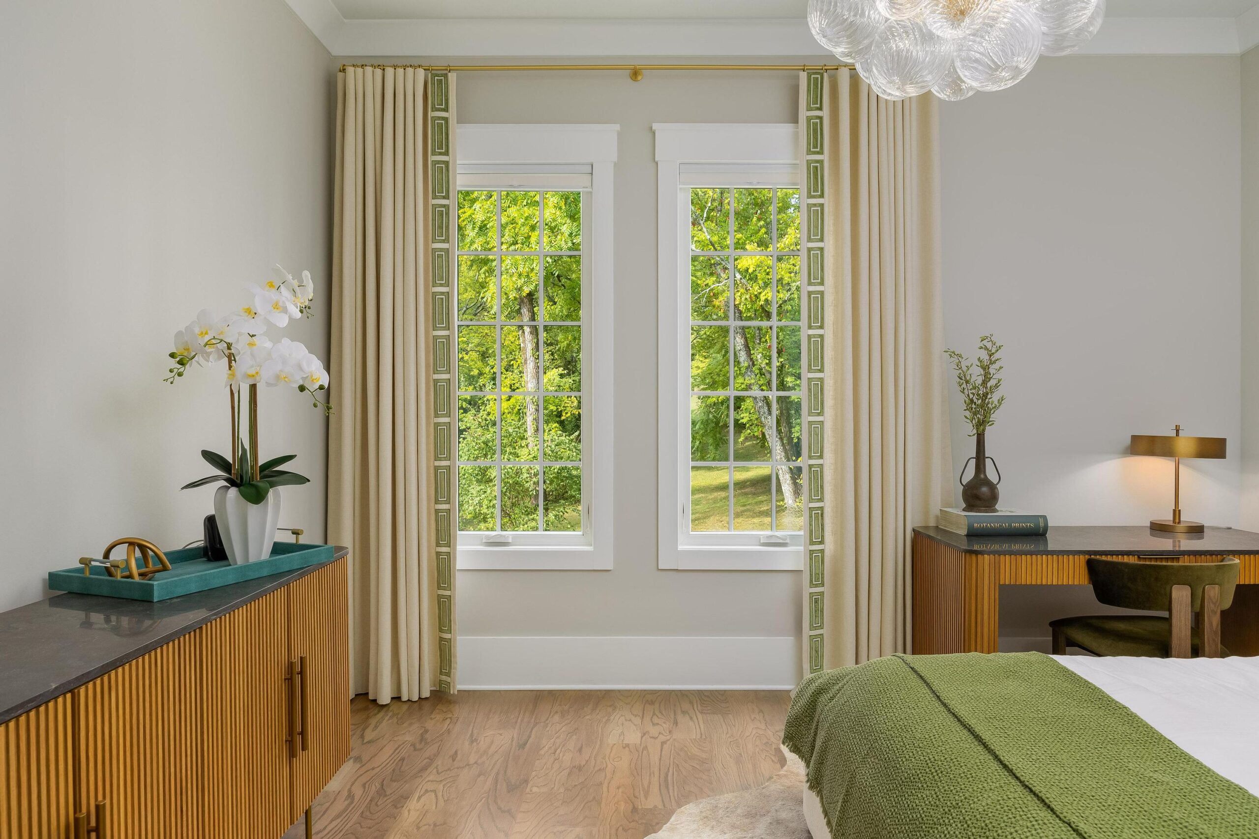A bright bedroom with large windows featuring elegant window treatments, light beige walls, wooden furniture, green accents, and a leafy outdoor view.
