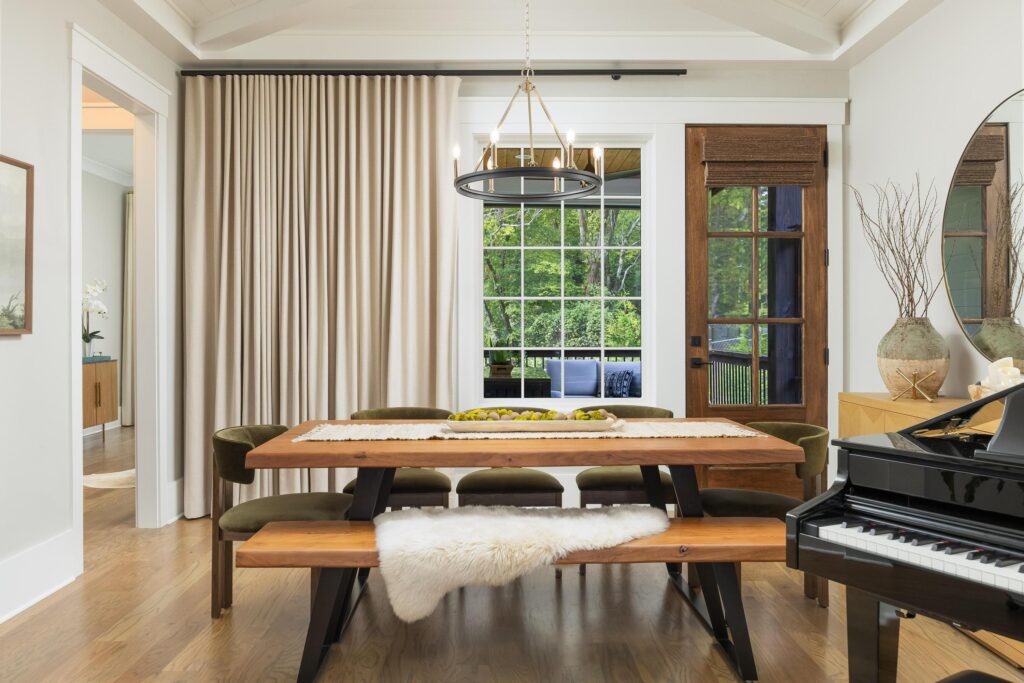 A modern dining room with a wooden table, benches, green chairs, a chandelier, piano, and large window dressed with elegant window treatment beside a glass door overlooking greenery.
