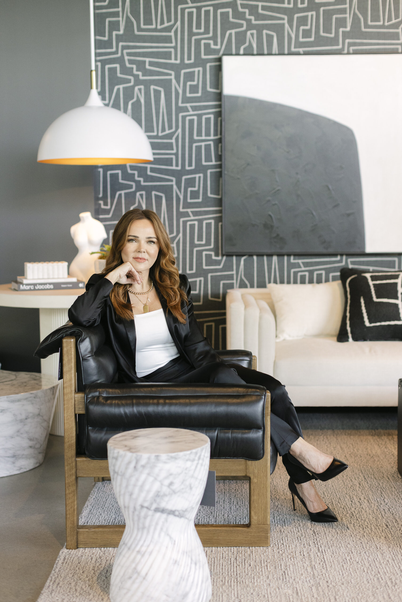 Woman in a black suit sits in a modern living room, where bold geometric wallpaper, abstract art, and marble accent tables showcase refined interior design.