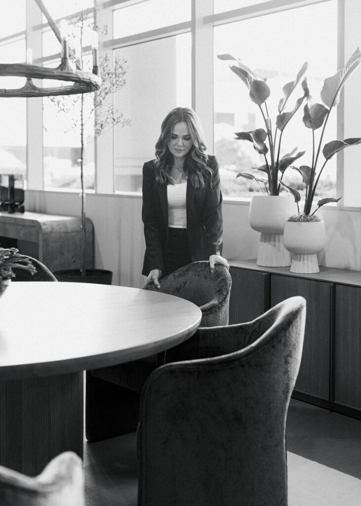 A woman in business attire stands behind a chair in a modern, well-lit office with large windows, potted plants, and stylish interior design.