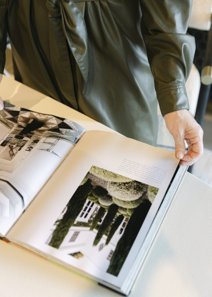 A person in a green long-sleeve garment is standing at a table, looking at an open book displaying photographs of architecture, gardens, and interior design.