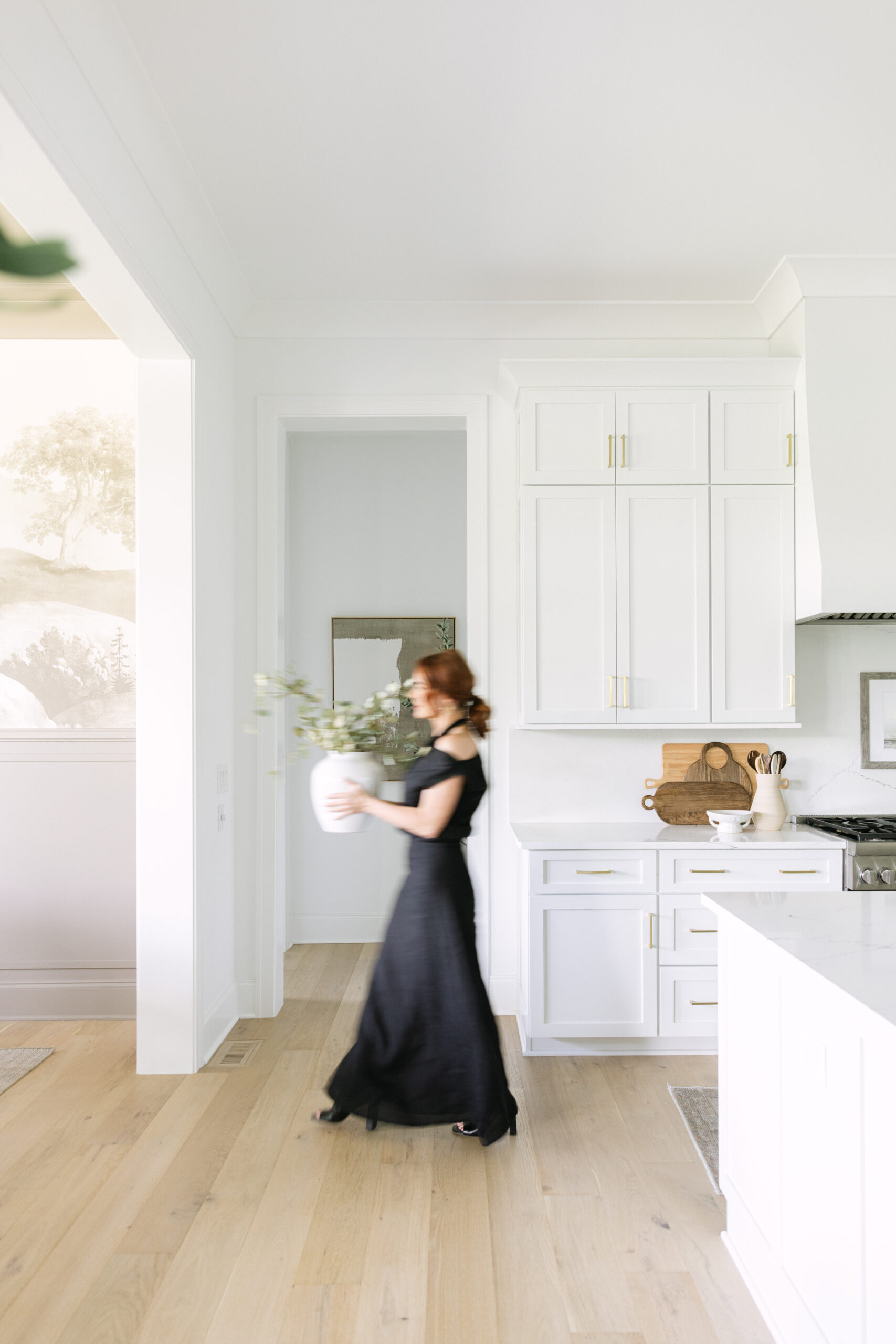 A woman in a black dress walks through a bright, modern home kitchen holding a vase with green branches. The kitchen features white cabinets and light wood flooring.