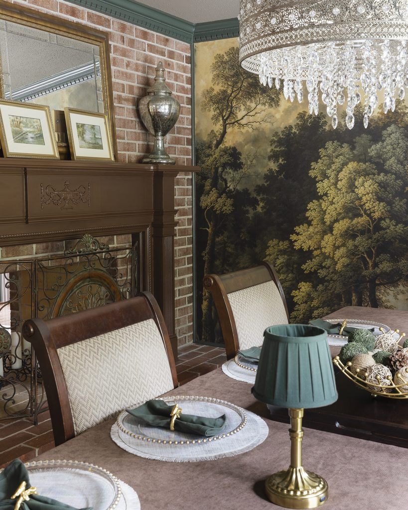 Elegant dining room with a brick fireplace, ornate mantel, crystal chandelier, and a mural of a forest scene. The room features a table set with green lamps, plates, and tulip-folded napkins.