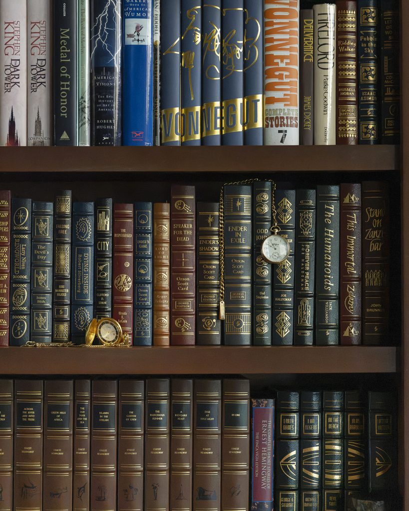 A bookshelf filled with classic and modern hardcover books, some with gold embossing, stands as a centerpiece in the room, while two small pocket watches rest elegantly on the shelves.