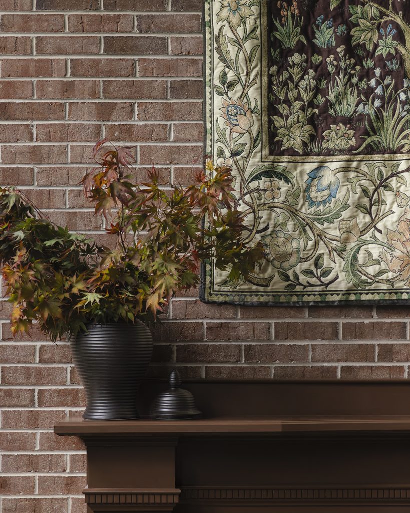 A dark vase with leafy branches sits on a brown mantel against a brick wall, adding charm to the room, with a floral embroidered tapestry hanging above.