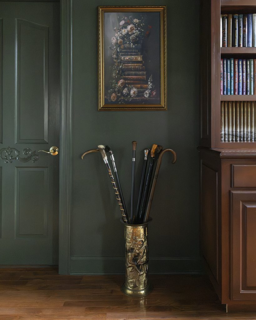 A brass umbrella stand with several canes is placed against a dark green wall beneath a framed floral painting, accenting the entrance to the room next to a door and a bookcase.