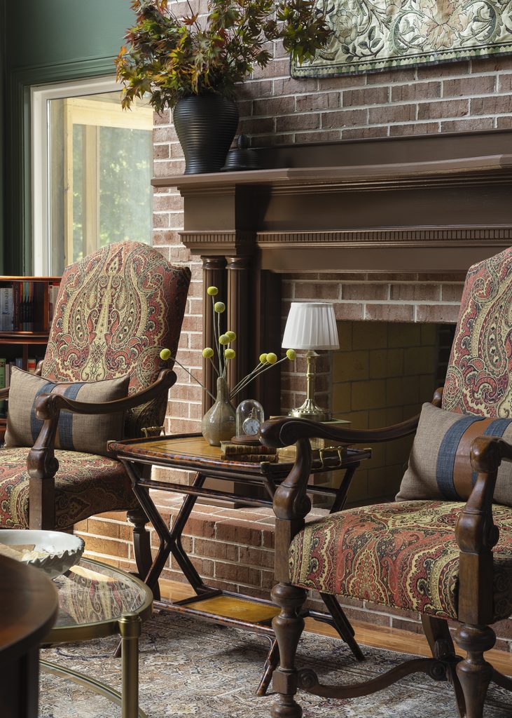 Two patterned armchairs sit by a brick fireplace in this inviting room, with a small table between them holding a vase, books, and a lamp. A decorative vase with branches is on the mantel above.