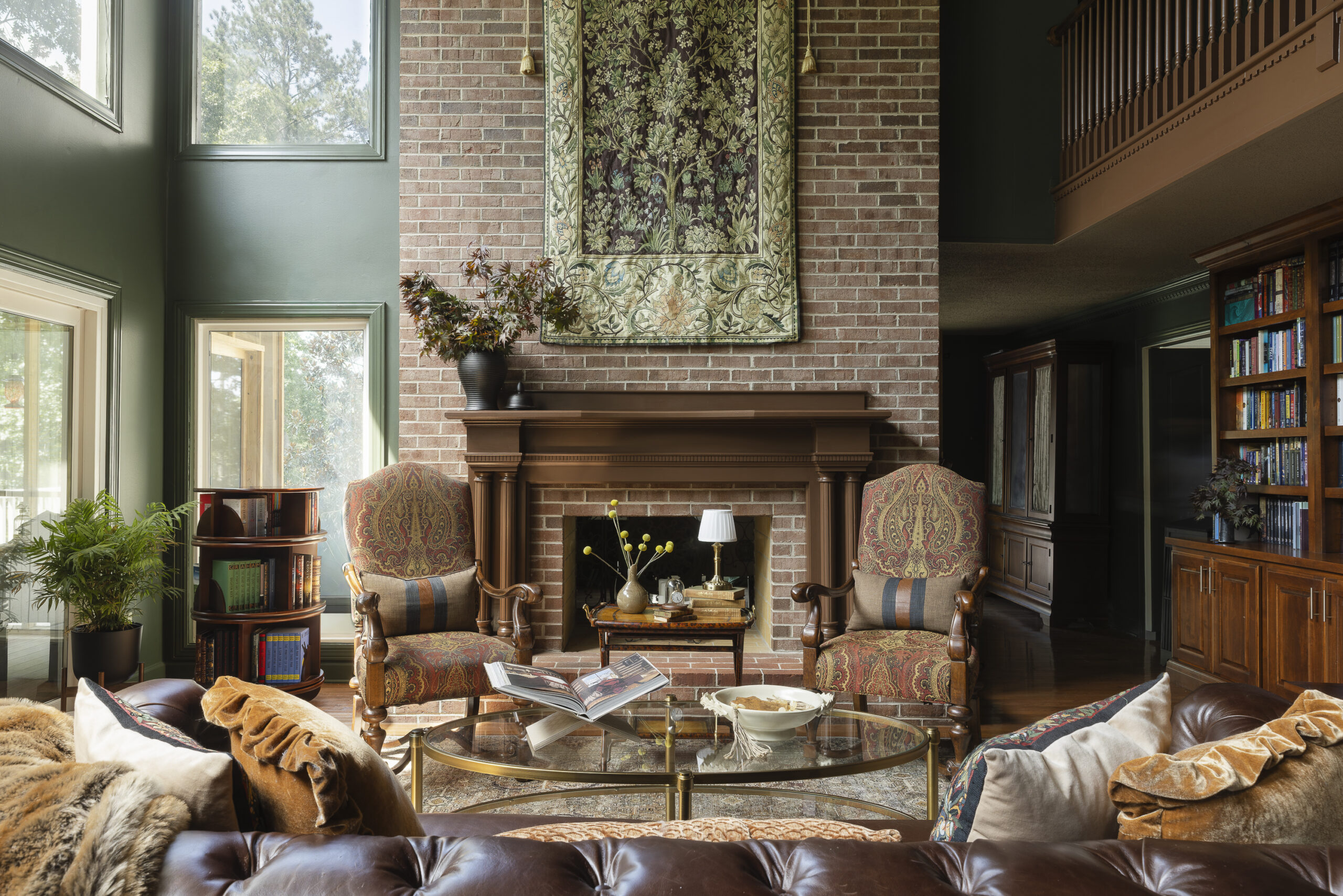 Traditional living room with a brick fireplace, ornate chairs, a tapestry above the mantel, bookshelves, and a glass coffee table with books and decorative items create a warm and inviting room.