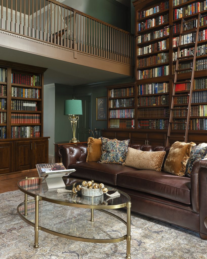 A leather sofa with patterned pillows sits in front of tall, filled bookshelves and a ladder in a cozy home library room. A glass coffee table holds a decor bowl and magazines.
