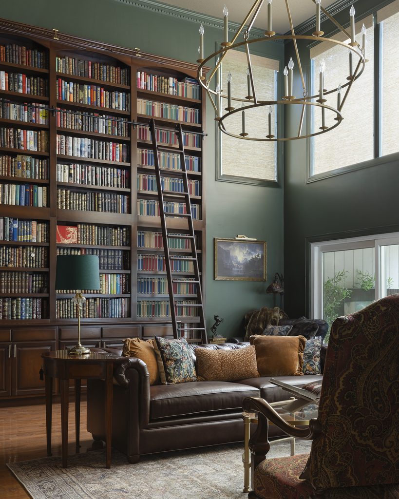 Home library room with tall bookshelves, a rolling ladder, leather sofa, patterned pillows, side table with lamp, chandelier, large windows, and framed painting on a green wall.