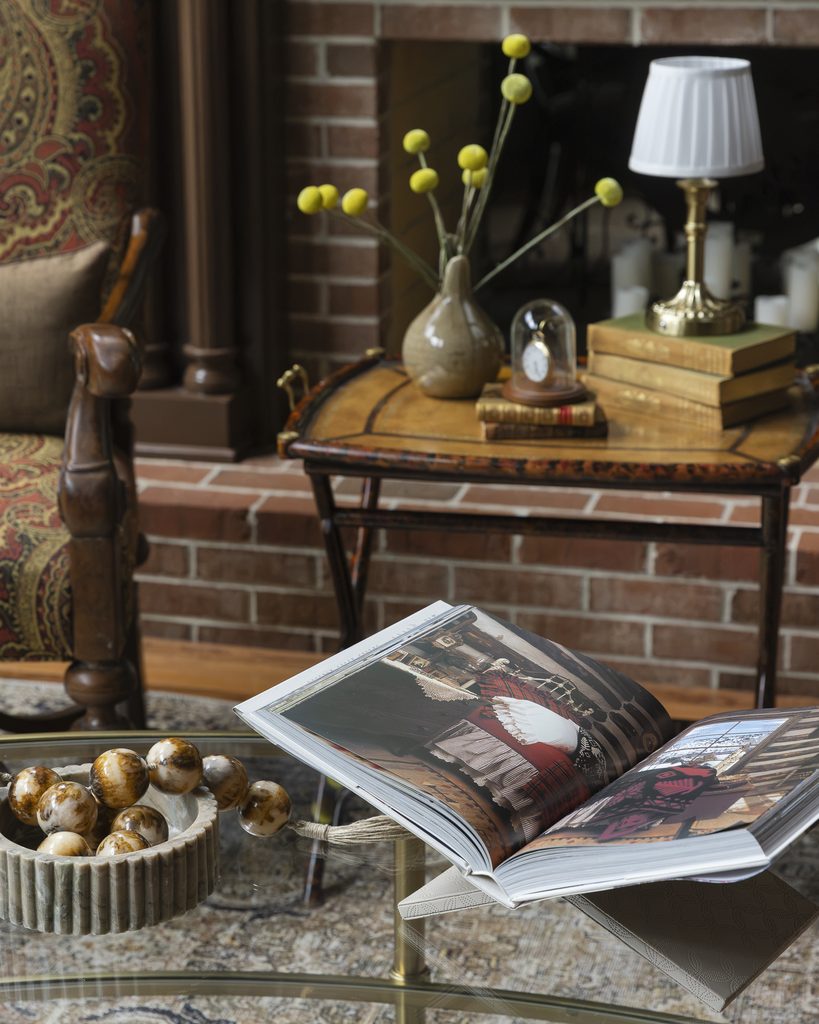 An open coffee table book displaying interior design photos of a room, resting on a glass table in front of a fireplace.