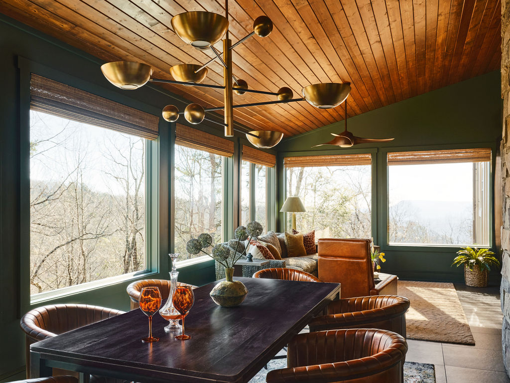 A dining room with a dark wood table surrounded by tan leather chairs under a large brass chandelier, with large windows overlooking a forest.