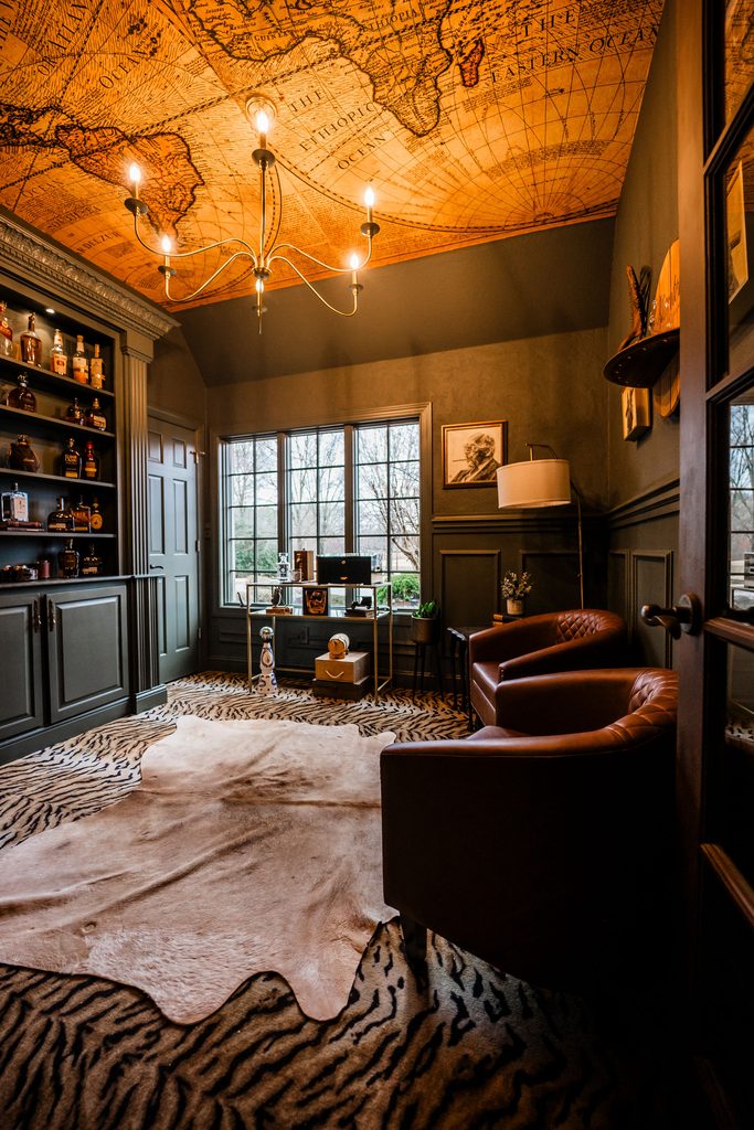 A study room with dark walls, two leather armchairs, a built-in liquor cabinet, and a map of the world on the ceiling.